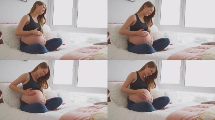 孕妇穿着健身服坐在家里的床上抱着肚子感觉婴儿踢