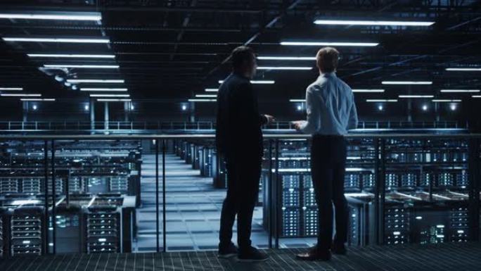 数据中心IT专家和电子商务企业家交谈，使用笔记本电脑。两位信息技术专业人士走在桥上俯瞰云计算服务器场