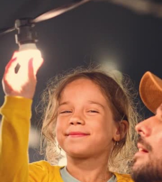 垂直屏幕: 父亲帮助他美丽的小女儿在家里的仙女灯后院安装了灯泡。父女击掌并庆祝成功修复。