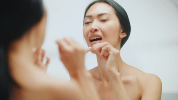 美丽的亚洲女人的肖像用牙线清洁她的天然洁白的牙齿，微笑在浴室的镜子里看着相机。晨间常规，牙齿卫生，健