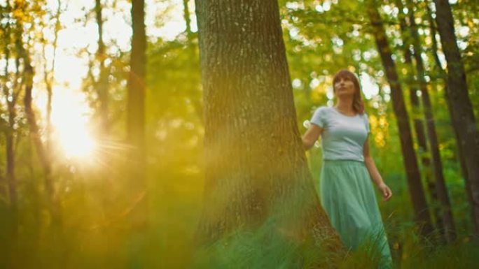 日落时分，穿着白色长裙的SLO MO女人在森林里散步时碰到树干