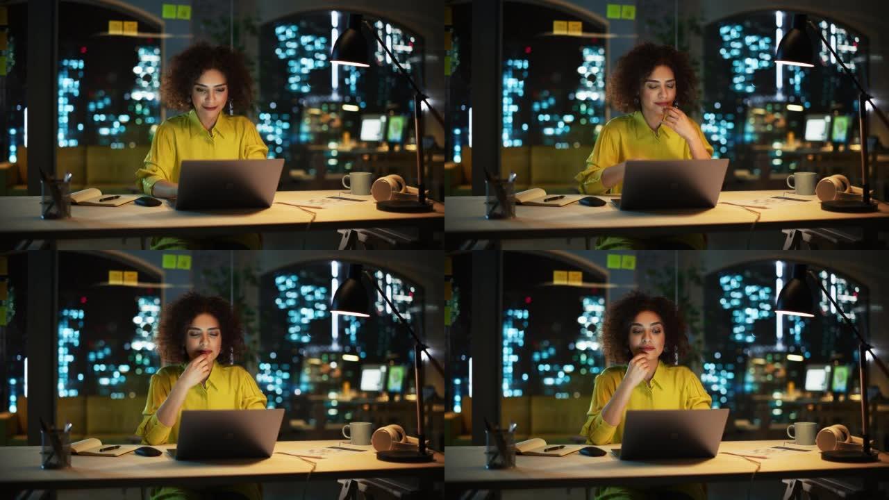 一个快乐的中东经理坐在创意办公室的办公桌前的肖像。时尚的女性，卷发，使用笔记本电脑，在夜间考虑营销机