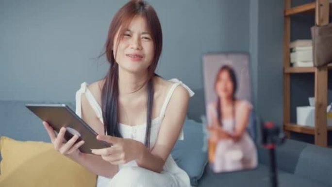 快乐年轻的亚洲女孩博客在手机摄像头前使用平板电脑在家里的客厅享受与追随者的问题答案。博客活动生活方式