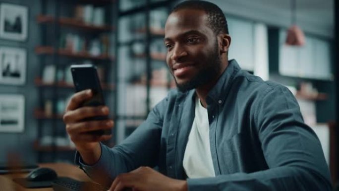坐在客厅的桌子旁，兴奋的黑人非洲裔美国人使用智能手机。快乐的男人在家里微笑，通过互联网与同事和客户聊