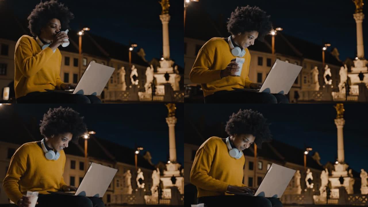 快乐的年轻女子坐在城市的长凳上，一边用笔记本电脑工作，一边喝咖啡