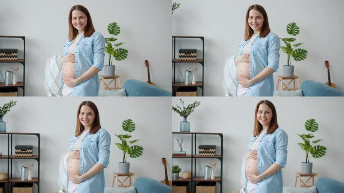 美丽孕妇的肖像微笑着看着相机抚摸着肚子在家
