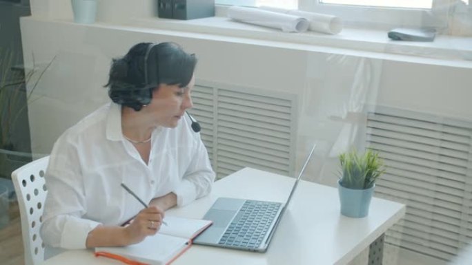 认真的女员工戴着耳机看着笔记本电脑屏幕并在办公室写字