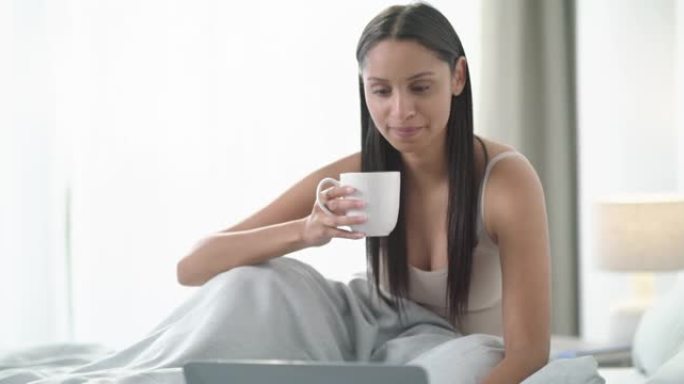 4k视频片段，一名年轻女子使用笔记本电脑并在家中床上喝咖啡