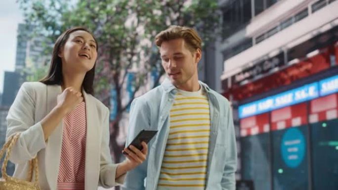 年轻时尚的多民族夫妇在大城市的街道上随意行走。迷人的日本女性向英俊的高加索男性展示智能手机。不同的朋