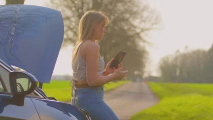 在乡间小路上沮丧的女人站在抛锚的汽车旁边，引擎盖朝上，用手机呼救-慢动作拍摄
