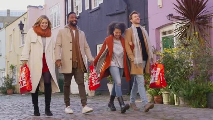 两对多元文化的夫妇在秋天或冬天参观城市时，手挽手走在鹅卵石的mews街上，带着出售购物袋-慢动作拍摄