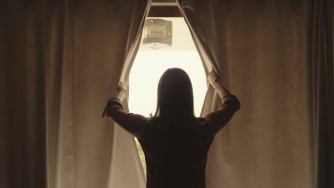 一个成熟的女人在家里放松的早晨打开卧室的窗帘。从新的一天开始，一位穿着剪影的高级女士的后视图看着窗外