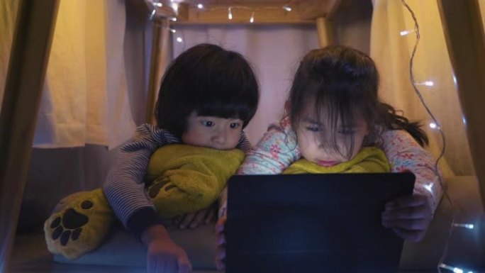 在自制床单下的女孩和男孩作为帐篷看着数字平板电脑。