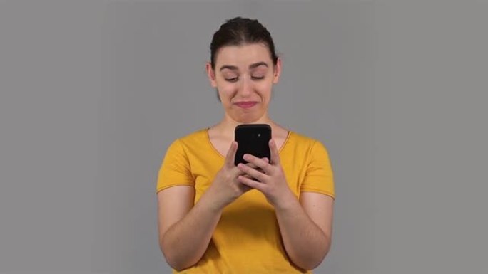 在灰色的背景下，身着黄色衬衫的年轻女子正开心地看着手机。社交媒体的概念