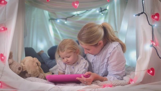 母亲和年幼的女儿在家里孩子卧室的自制营地里看着数字平板电脑