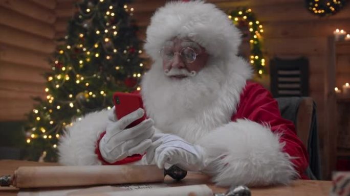 圣诞老人在他装饰精美的小屋中，在他的红色手机上进行视频聊天，愉快地道别