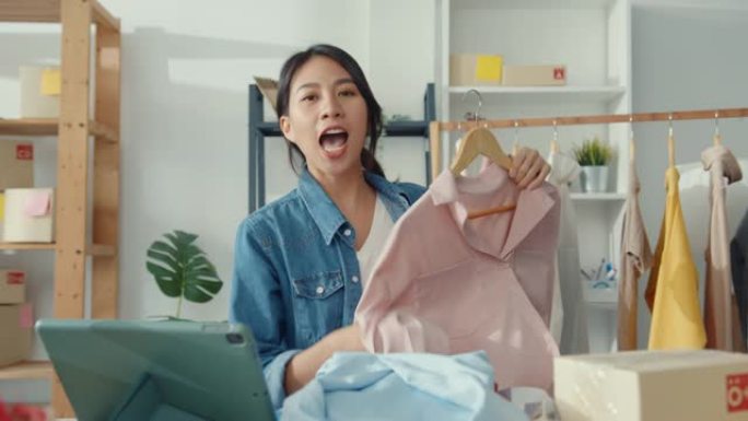 年轻的亚洲女士时装设计师使用手机接收订单，并在商店在线展示服装录制视频直播。