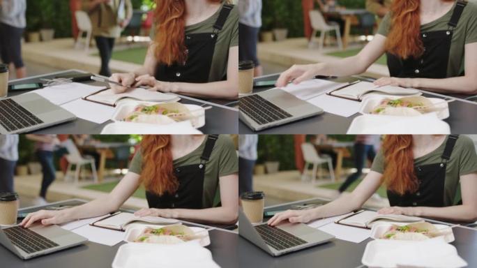 女人在午餐时拿着笔记本电脑，在笔记本上写字，在创意工作场所工作。在早餐时记下报告、在线营销研究和一个