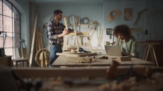 两个木匠在木制品车间一起工作。多元文化的男女同事看着笔记本电脑上的蓝图，创造了一种新的椅子设计。