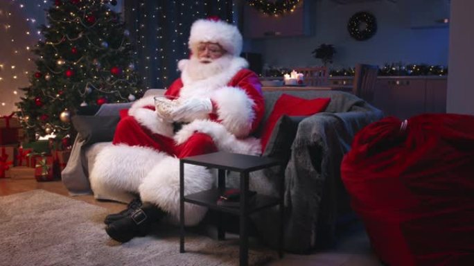 圣诞老人坐在装饰精美的客厅里过圣诞节，坐在灰色的沙发上看电视，吃着爆米花。圣诞老人在情感上担心电视上