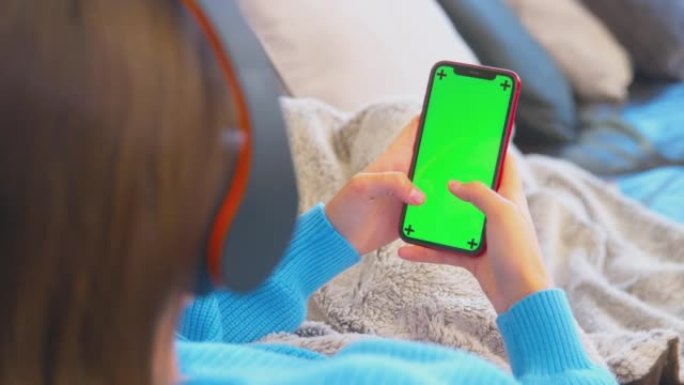 十几岁的女孩躺在沙发上戴着无线耳机从绿屏手机流内容-慢动作拍摄