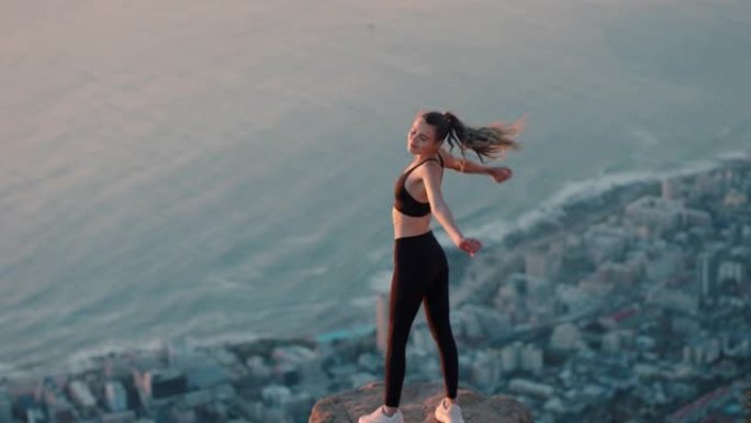 年轻女子举起手臂在山顶庆祝成就悬崖边缘的女孩看着日落时美丽的景色享受旅行冒险