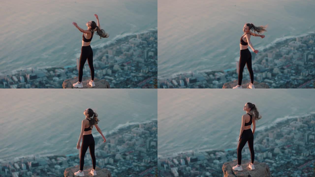 年轻女子举起手臂在山顶庆祝成就悬崖边缘的女孩看着日落时美丽的景色享受旅行冒险