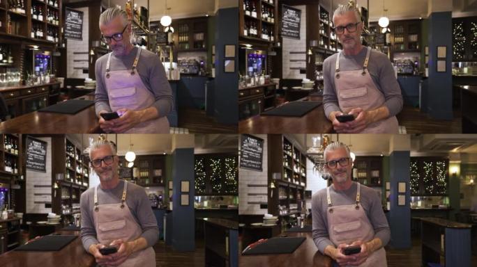 英俊的酒吧老板靠在酒吧柜台上的视频肖像在智能手机上发短信，然后面对相机微笑