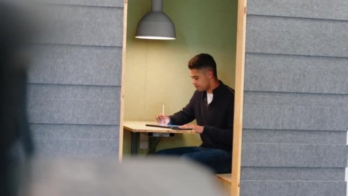 年轻人坐在小隔间里，用手机在数字平板电脑上做笔记