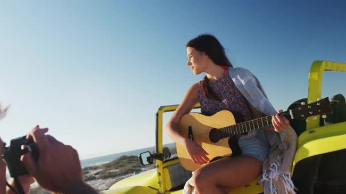 白人男子为坐在海边的沙滩车里弹吉他的女人拍照