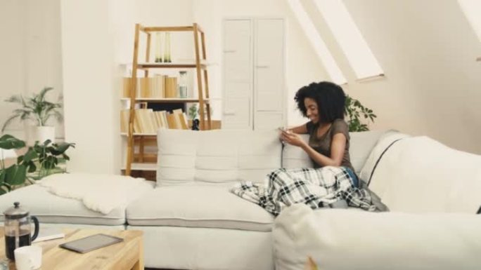 一名年轻的非洲裔美国妇女坐在家里的沙发上使用手机。一位美丽的混血女性，非洲裔在客厅的沙发上放松时上网