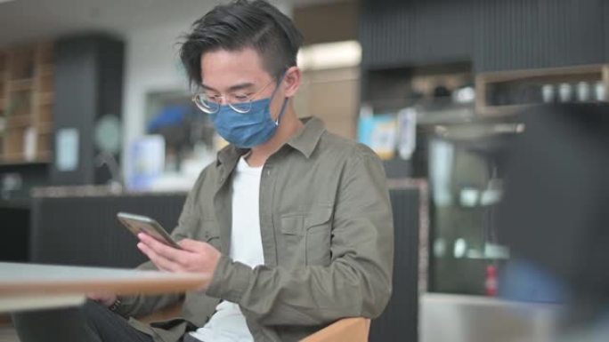 戴着防护口罩的亚洲中国少年男孩坐在咖啡馆里滚动并查看社交媒体