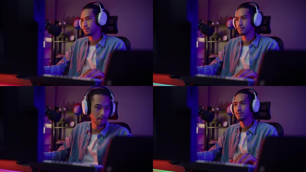 快乐亚洲男玩家戴着耳机参与玩视频游戏彩色霓虹灯电脑，晚上在客厅的平板电脑上录制播客现代房子。