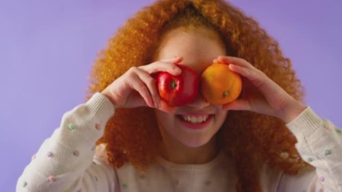 在紫色工作室背景下，红发年轻女孩拿着苹果和橙色像眼镜一样在脸前拍摄-慢动作拍摄