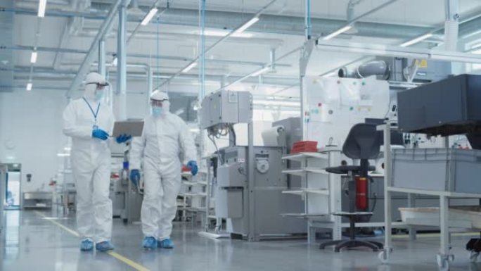 两个穿着无菌工作服的科学家带着笔记本电脑走在重工业工厂，检查工业数控机床设置和配置生产功能。