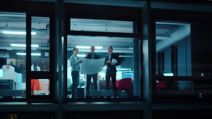 窗外的镜头: 戴着安全帽的建筑师和工程师站在办公室的窗户旁边，看着纸上的蓝图。高级专家向他的学徒解释