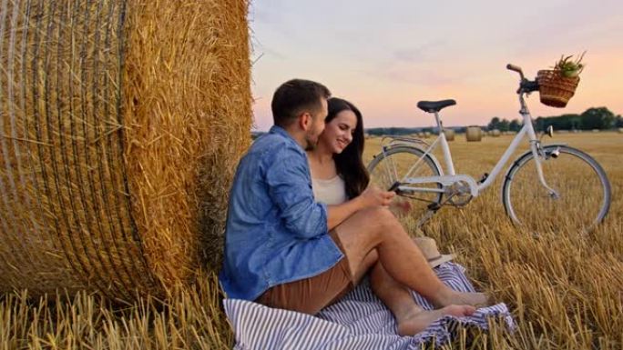 慢动作，一对成年夫妇在日落时坐在一吨重的草捆旁边看着电话的手持镜头