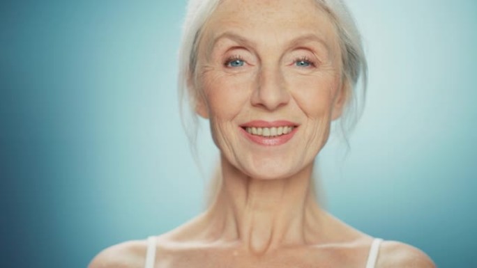美丽的高级女人转身，看着相机，笑容灿烂。漂亮的老年女性，有着灰色的头发和蓝色的眼睛的自然美。抽象蓝色