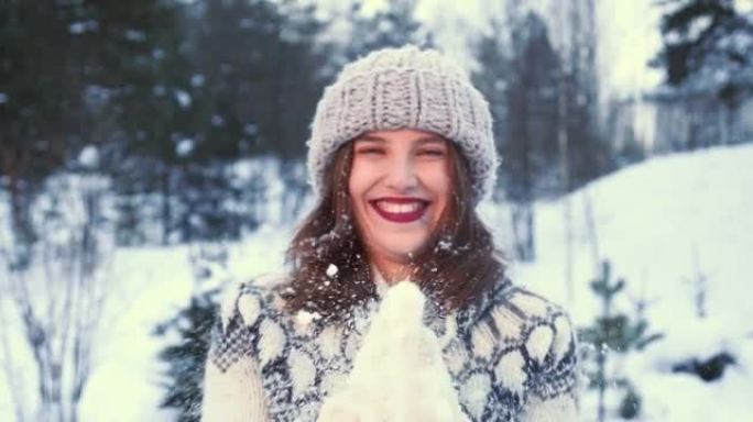 寒假。美丽的年轻幸福的女人戴着帽子，手套对着镜头吹雪，对着森林慢动作微笑