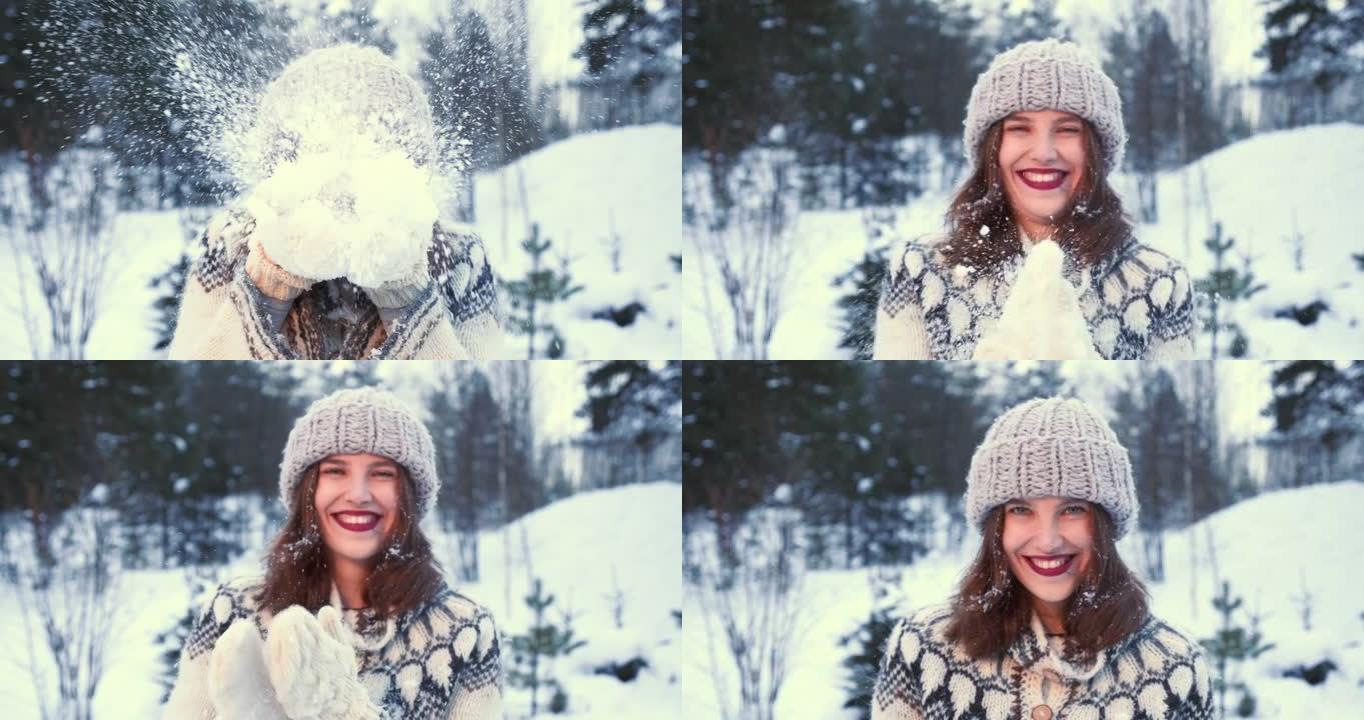 寒假。美丽的年轻幸福的女人戴着帽子，手套对着镜头吹雪，对着森林慢动作微笑