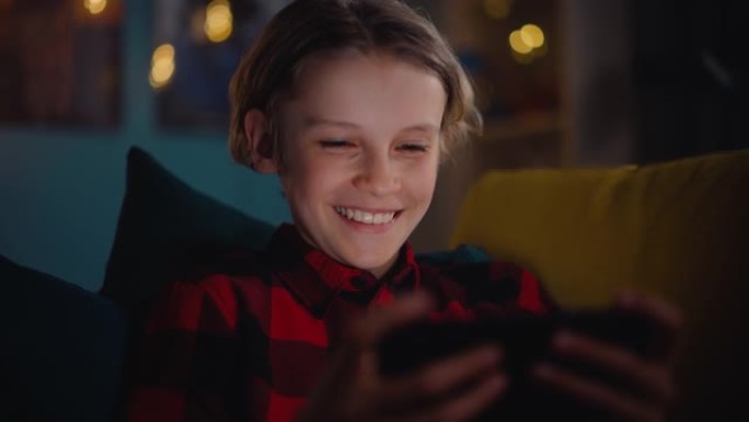 可爱的小男孩坐在黑暗舒适的房间的沙发上，在家使用智能手机。快乐的青少年浏览有趣的在线内容，在社交媒体