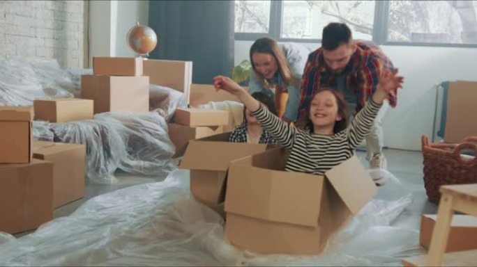 年轻有趣快乐的父母推动兴奋可爱的孩子在地板上的纸箱庆祝搬进新公寓。