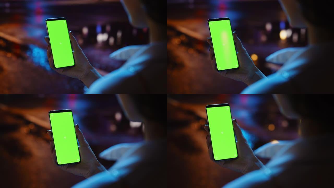 美丽的女人在充满霓虹灯的夜晚城市街道上行走时使用色度键智能手机。女性使用绿屏手机。肩以上静态中特写镜