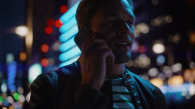 英俊的男人在智能手机上聊天，穿过充满霓虹灯的夜晚城市街道。市区时尚男性接听手机的肖像。中枪