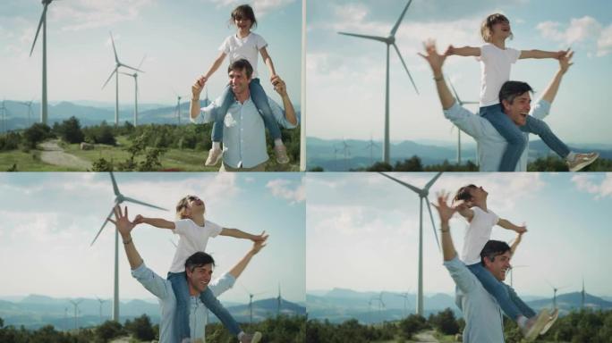 电影拍摄的快乐父亲在风电场中奔跑，笑着将女儿扛在肩上。父母和孩子在背景中带有风力涡轮机的绿色田野中玩