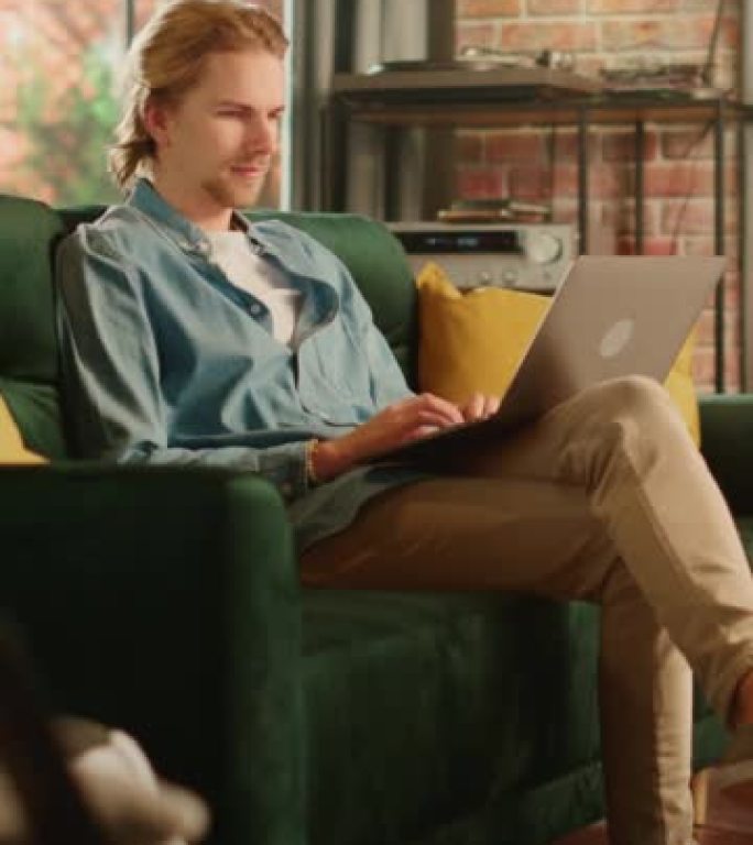 垂直屏幕: 年轻英俊的男人坐在阁楼公寓的沙发上，在家使用笔记本电脑工作。创意男性查看社交媒体，浏览互