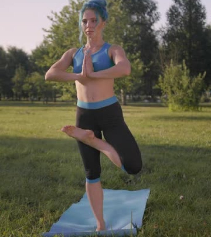 一个有吸引力的健身瑜伽老师的垂直视频，他穿着绿松石的瑜伽服装，穿着绿松石的头发，在公园的草地上做瑜伽