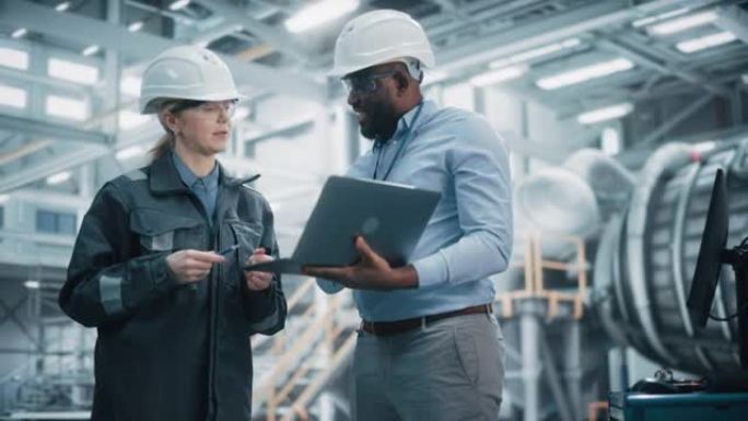 两名穿着安全制服和安全帽的专业重工业工程师在笔记本电脑上工作。非裔美国技术人员和女工在工厂开会。