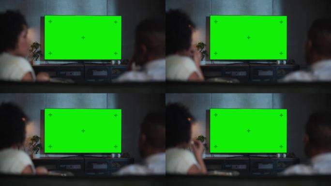 黑人夫妇的后视图在沙发上吃爆米花，并在阁楼房间看带有绿屏模拟显示的电视。Rack Out Focus