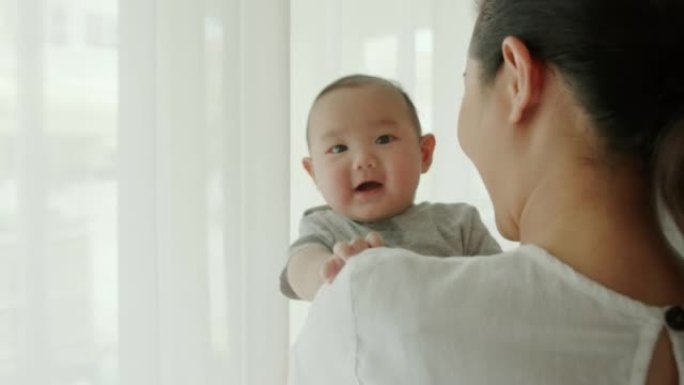 后视快乐而微笑的单身母亲背着可爱的新生男婴，早上看着窗外的客厅。